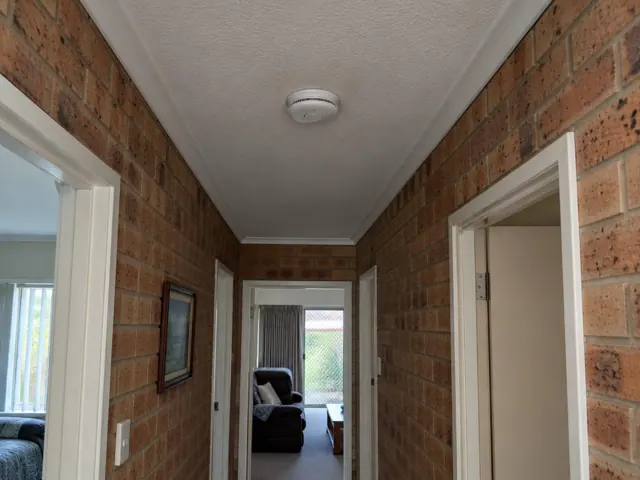 Ceiling-mounted smoke alarm in a brick hallway with doorways and carpeted room beyond in Labrador